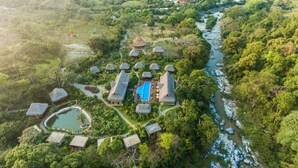 Vista aérea de um resort com edifícios de telhado de palha, piscina e rio.