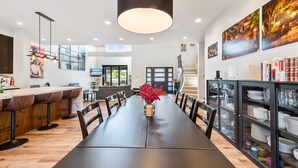 A modern dining area with a long table, chairs, a vase of red flowers, and a kitchen in the background. - 4BR Resort Near Beach/Disney; Pool is +$149 /night (Huntington Beach)