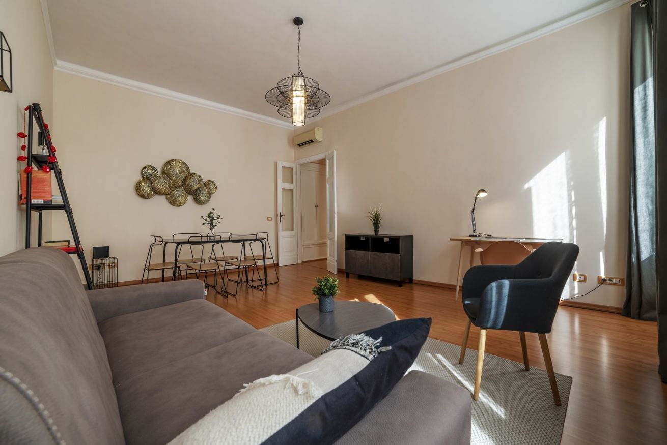 Een moderne woonkamer met een bank, een eettafel, een klein bijzettafeltje, een stoel, een lamp en een decoratief wandobject.
