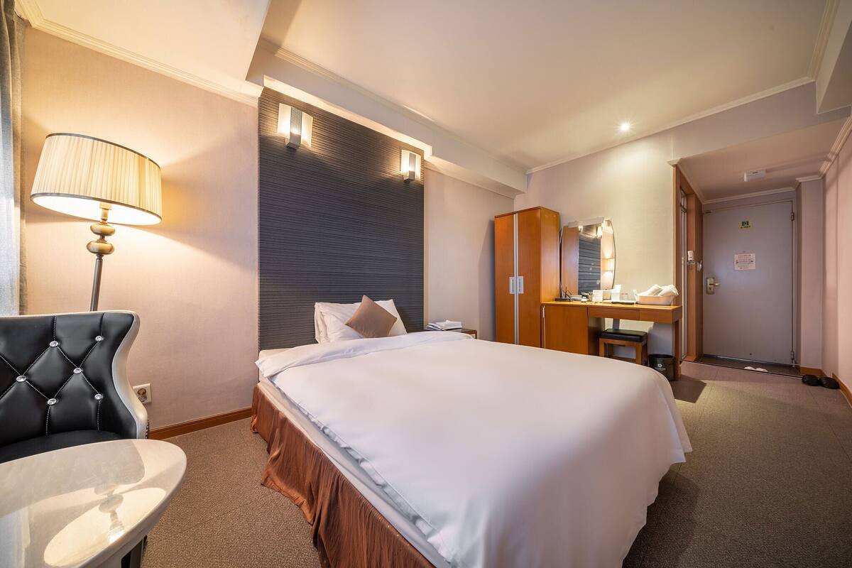 A hotel room with a bed, a desk, a chair, a lamp, a mirror, and a wardrobe.