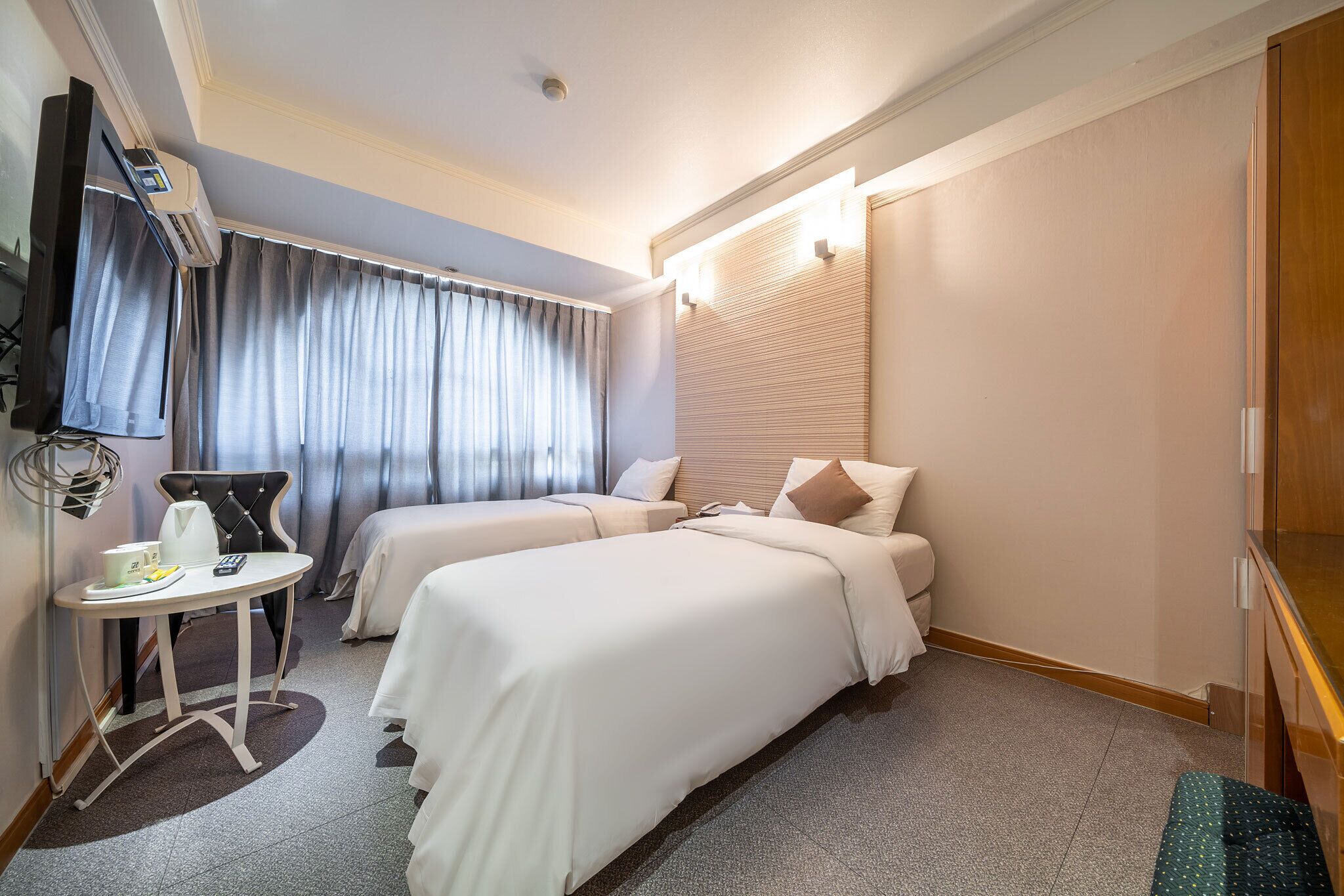Habitación de hotel con dos camas, una mesita con una tetera y una taza, un televisor y una ventana con cortinas.