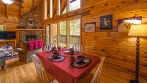 Un interior acogedor de cabaña con una mesa de comedor dispuesta para cuatro, paredes de madera, una chimenea de piedra y un televisor en una esquina.