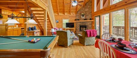 A wooden cabin interior with a pool table, seating area, fireplace, and large windows.