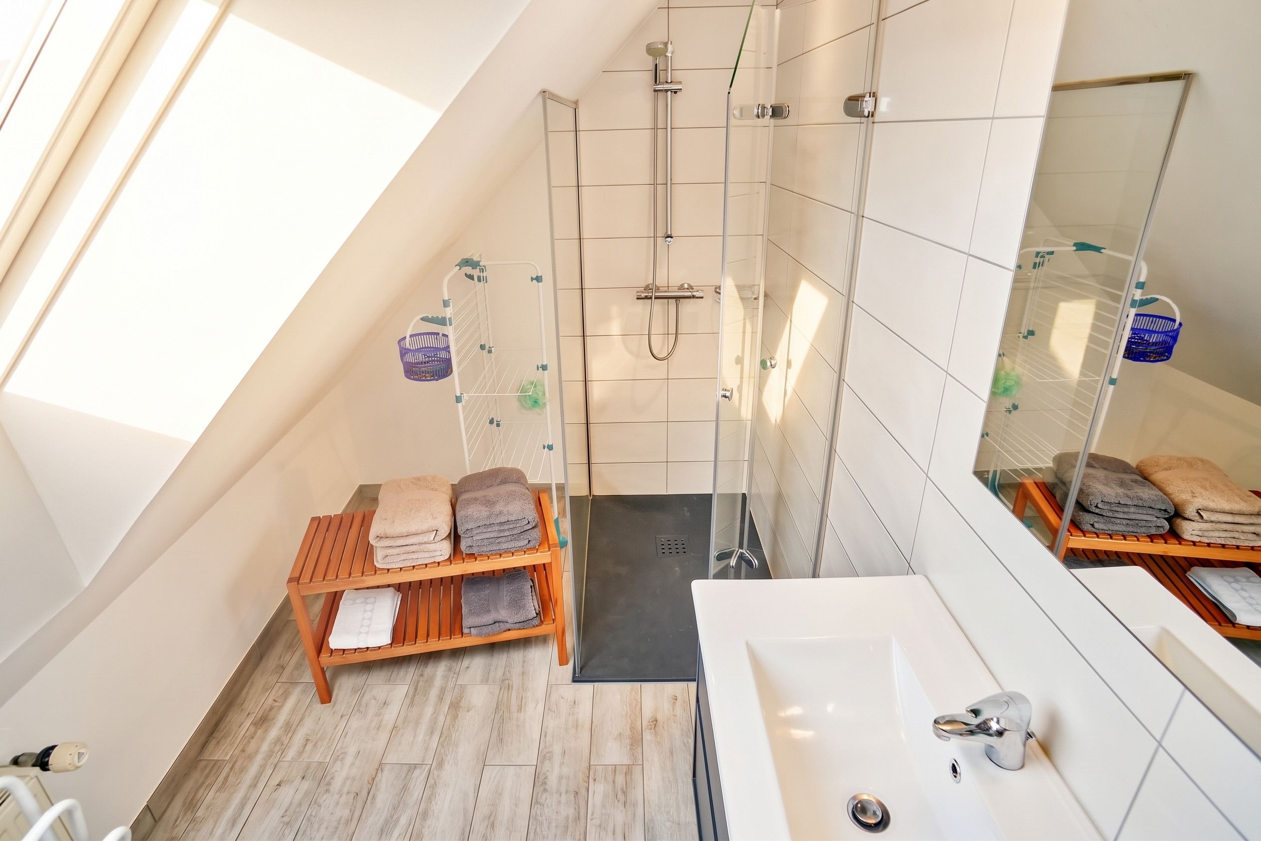 Ein modernes Badezimmer mit Dusche, einer Holzbank mit Handtüchern, einem großen Spiegel und einem Oberlicht.