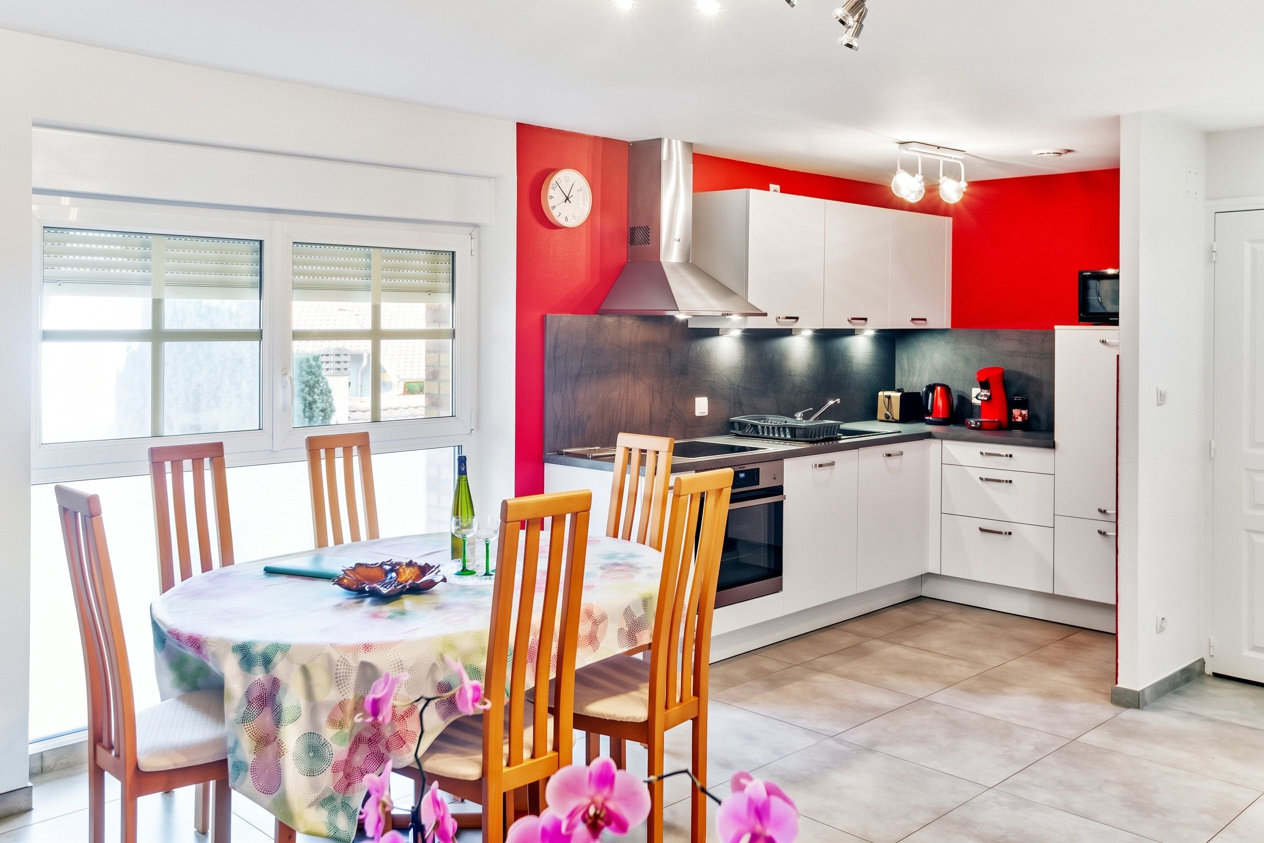 A modern kitchen with white cabinets, a red accent wall, a built-in stove, and a dining area with wooden chairs and a table.