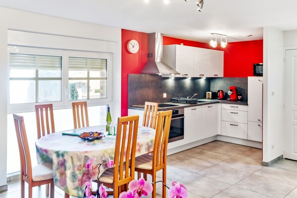 A modern kitchen with white cabinets, a red accent wall, a built-in stove, and a dining area with wooden chairs and a table.