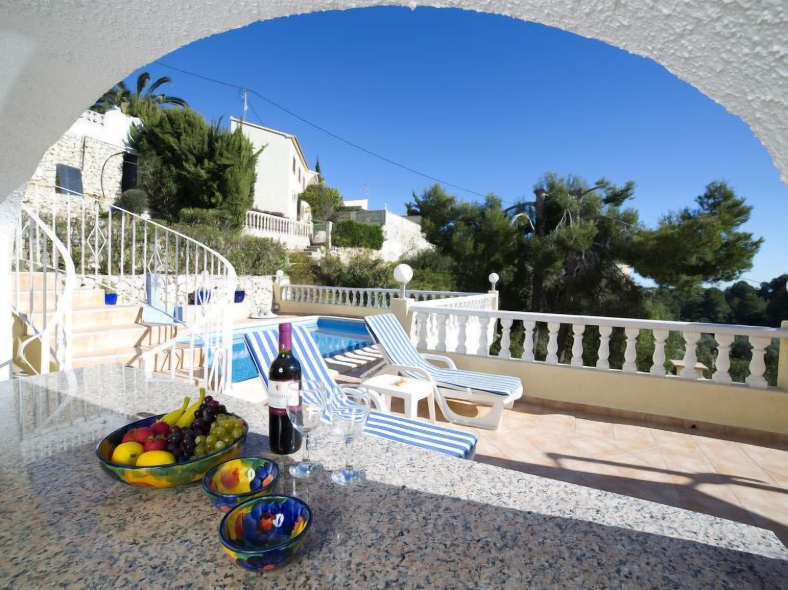 A terrace with a pool, lounge chairs, and a table with fruit bowls and a bottle of wine.