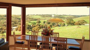 Un coin repas avec une table et des chaises, un vase de fleurs et une vue sur un paysage verdoyant, visible à travers de grandes fenêtres.