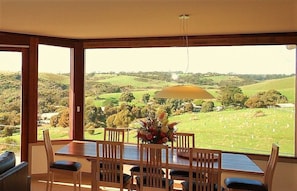 A dining area with a table and chairs, a vase of flowers, and a view of a green landscape through large windows.
