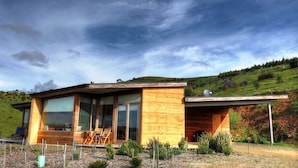 Une maison moderne avec de grandes fenêtres, une terrasse avec des chaises et une vue sur une colline verdoyante.