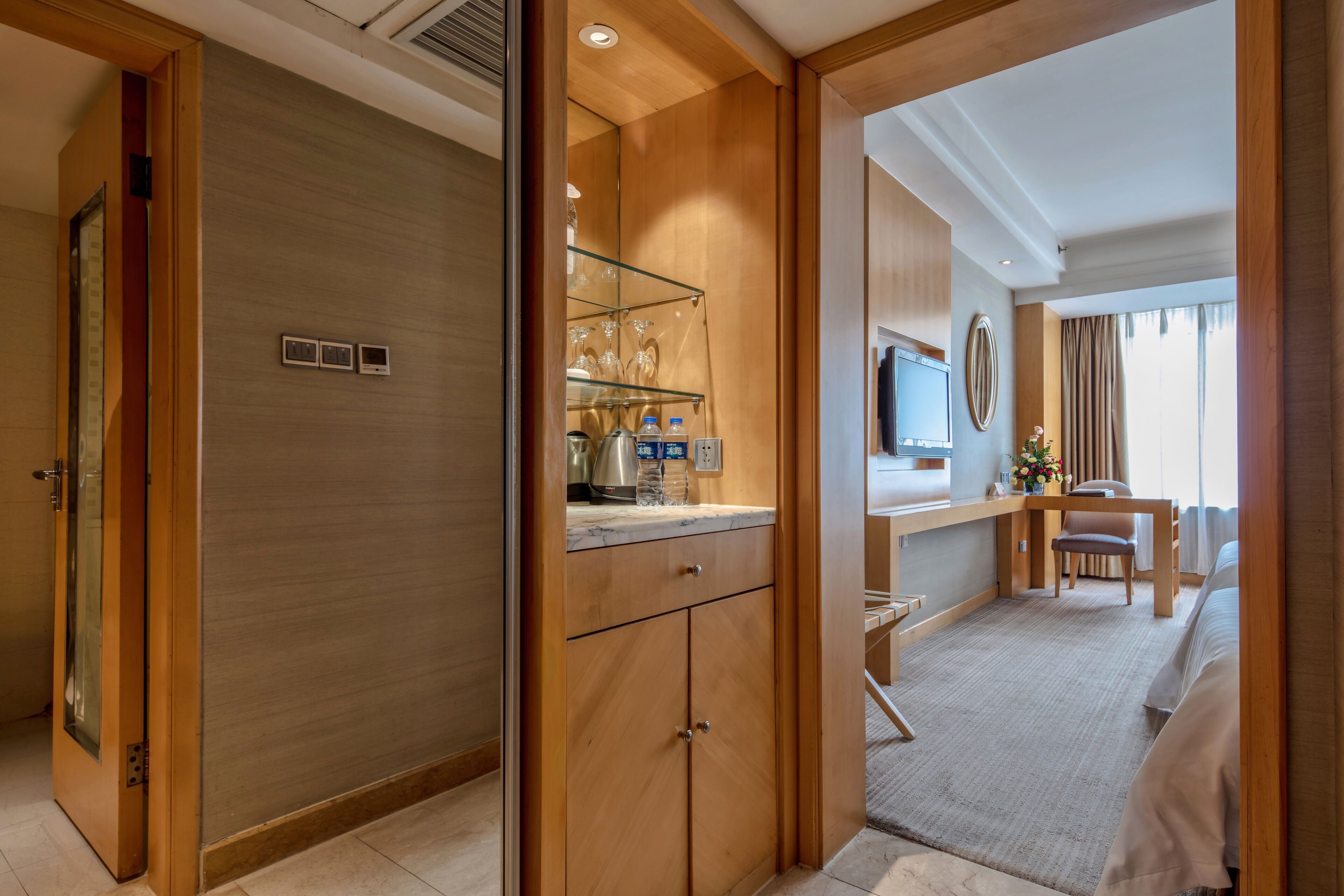 A hotel room with a wooden wardrobe, a glass shelf with toiletries, a desk with a chair, a bed, and a TV mounted on the wall.