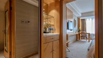 A hotel room with a wooden wardrobe, a glass shelf with toiletries, a desk with a chair, a bed, and a TV mounted on the wall.