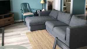 Uma sala de estar com paredes verde-água, um sofá modular cinza, um rack de madeira para TV, uma cadeira azul, um abajur e um relógio.