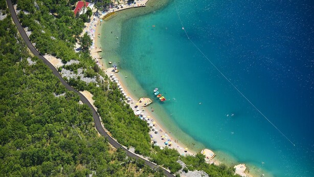An aerial view of a coastal area with a road curving through a forested area, leading to a beach with umbrellas and a small dock. - Villa Estrella (Dramalj)