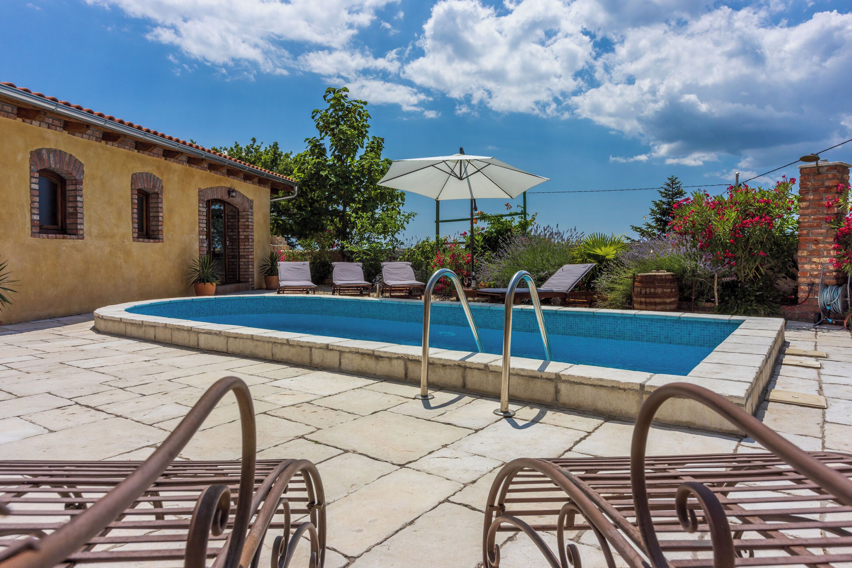 Ein Swimmingpool mit Liegestühlen und Sonnenschirm, umgeben von einem Backsteinhaus und Grünflächen.
