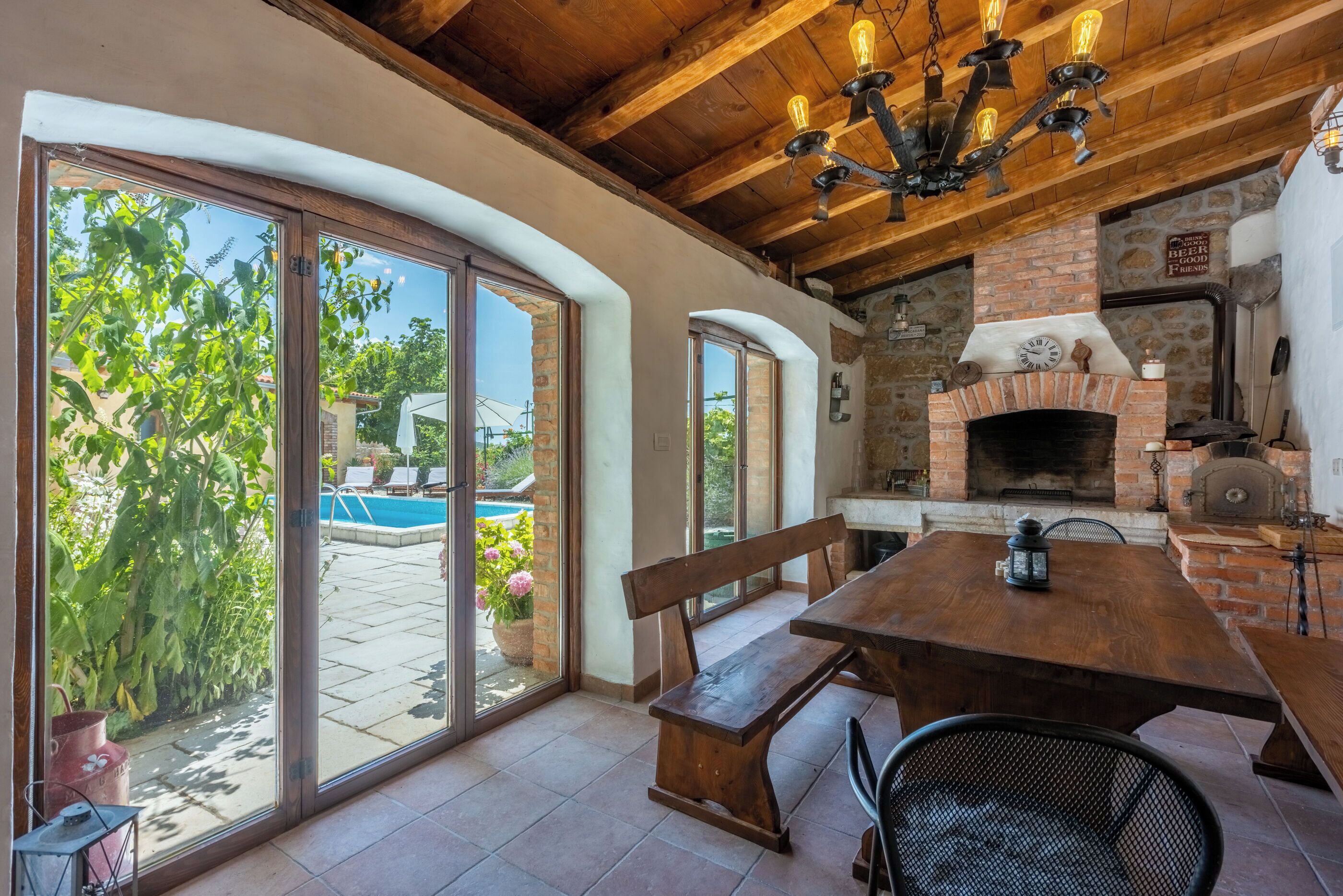 A patio with a wooden table and benches, a brick fireplace, and a pool in the background.