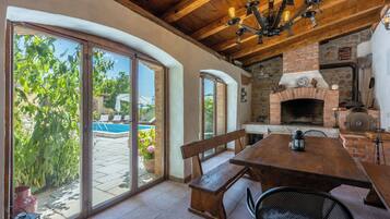 A patio with a wooden table and benches, a brick fireplace, and a pool in the background.
