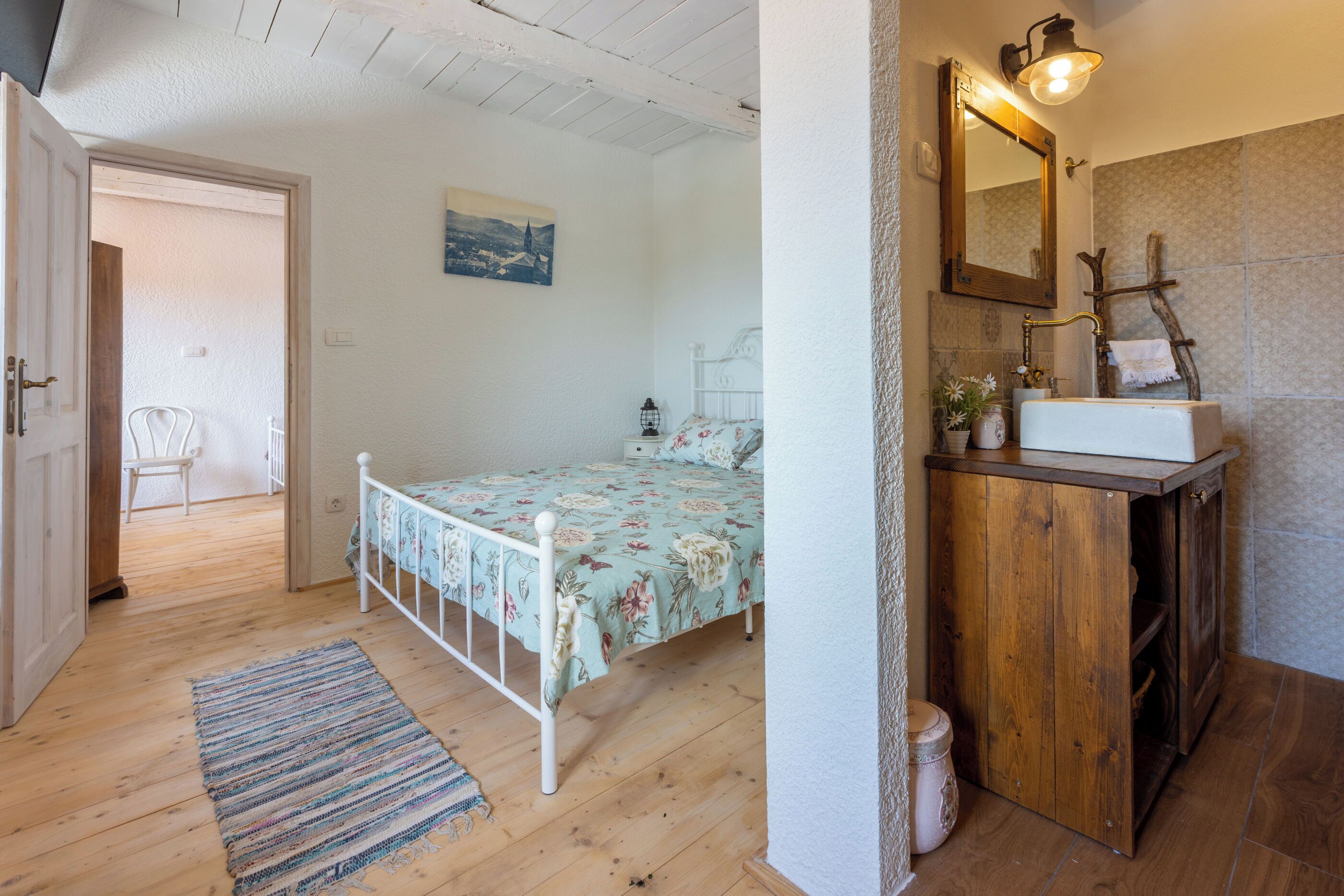 Un dormitorio con suelo de madera, una cama con ropa de cama floral, una mesita de noche, un armario de madera con espejo, y una puerta que da a otra habitación.