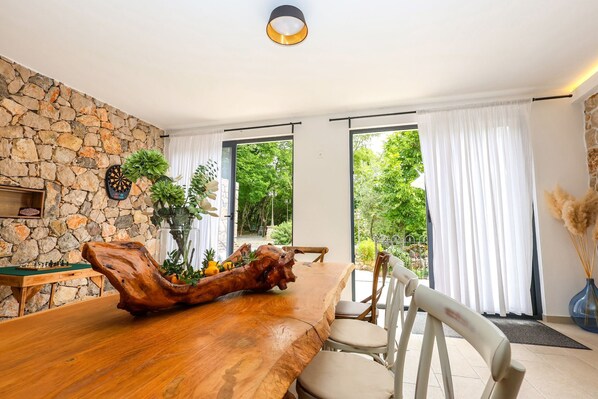A dining area with a wooden table, chairs, a stone wall, and large windows. - Villa Siesta (Grizane)