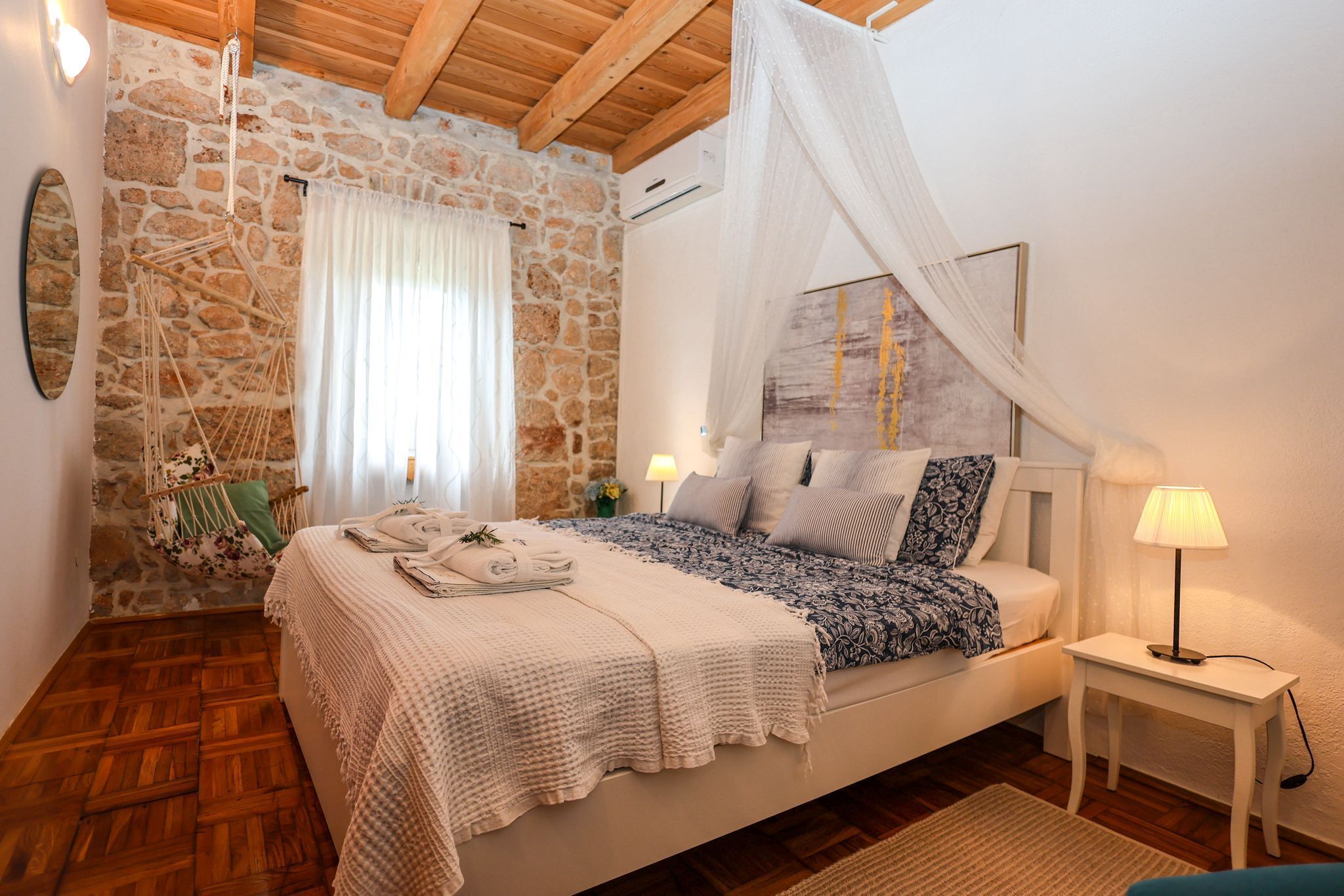 Un dormitorio con pared de piedra, suelo de madera, cama con ropa blanca, mesita de noche con lámpara y una silla colgante.