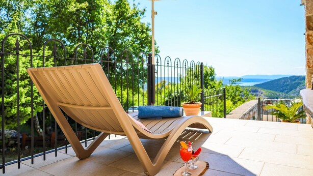 Un transat avec un coussin bleu et une boisson, sur une terrasse offrant une vue sur la verdure et les collines.