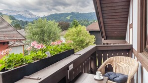 Een balkon met een rieten stoel, een klein tafeltje met een kopje en een bloembak met roze bloemen.