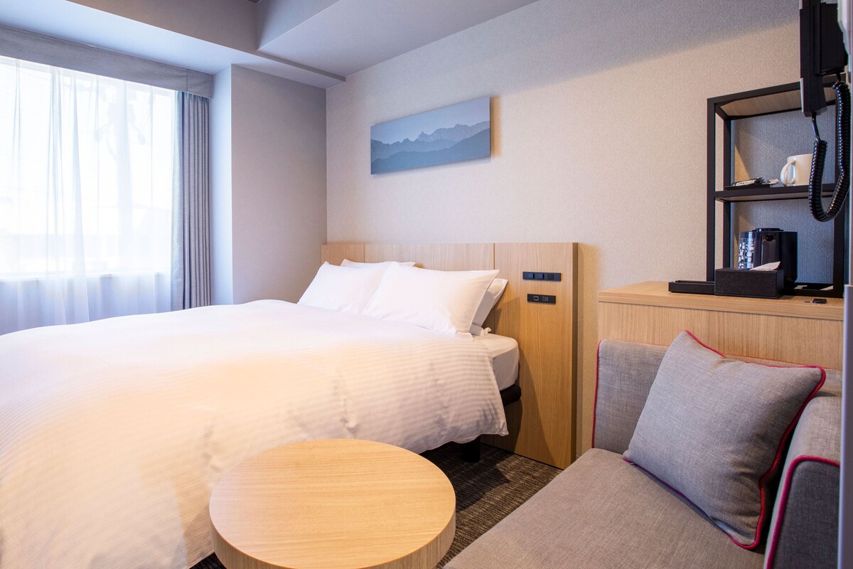 Ein Hotelzimmer mit einem großen Bett, einem kleinen runden Tisch, einer Sitzecke und einem Minikühlschrank. An der Wand hängt ein Gemälde.