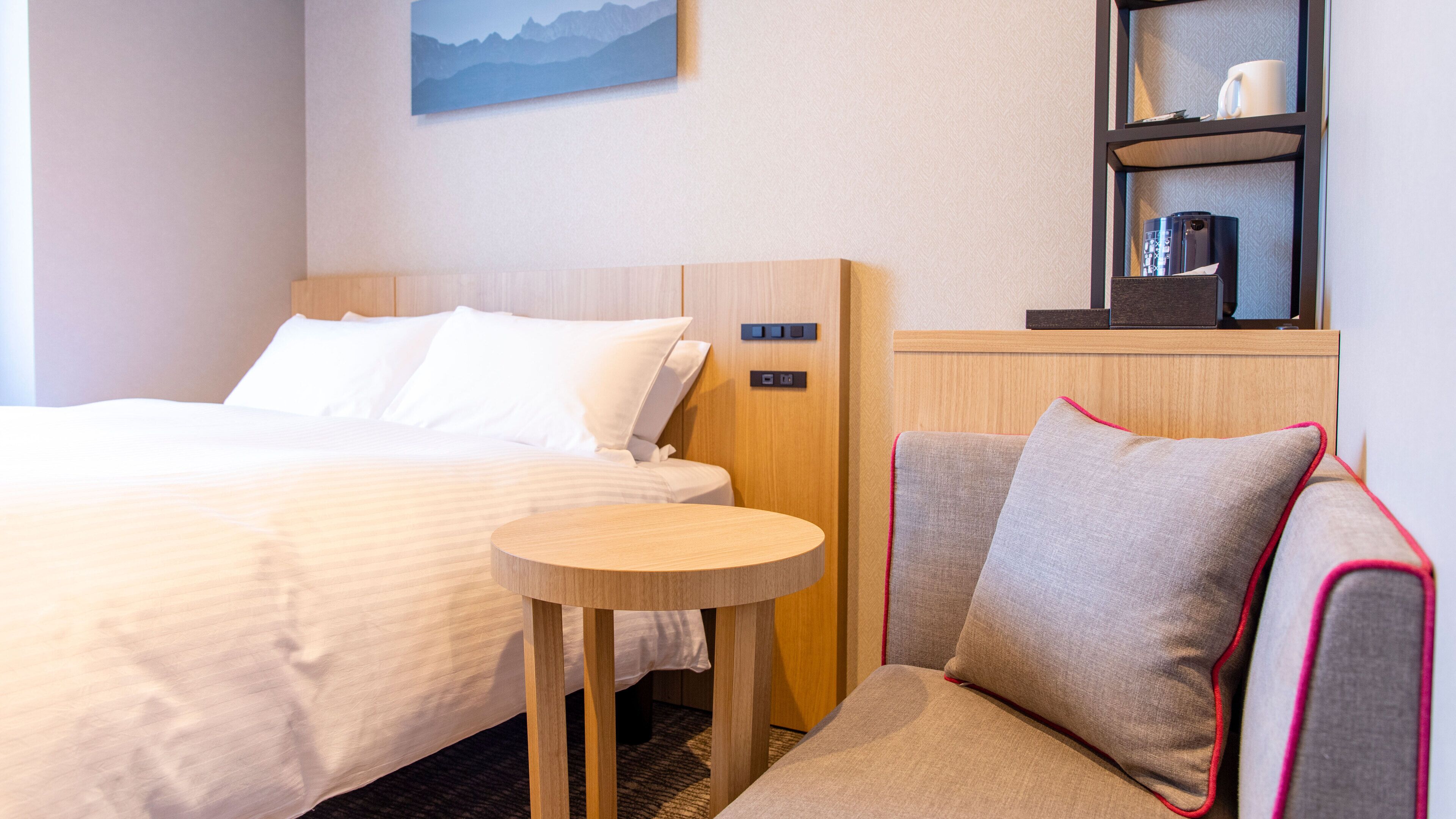 Une chambre d’hôtel avec un lit, une table ronde en bois, un canapé avec un coussin et une étagère sur laquelle se trouvent une machine à café et une tasse.