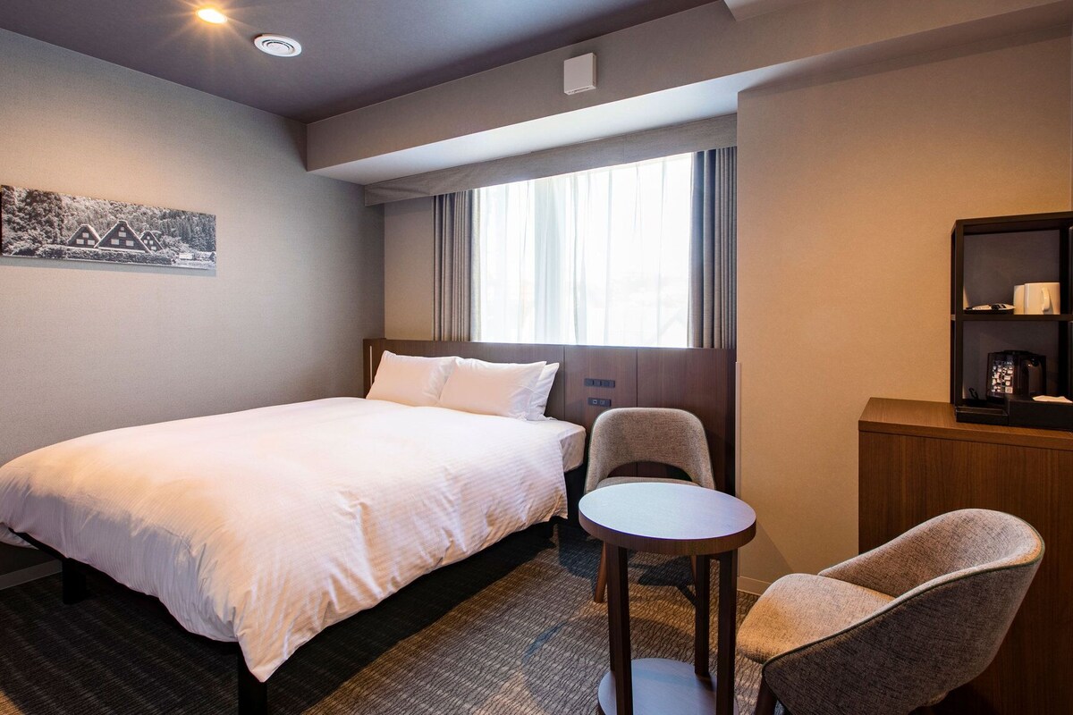 Ein Hotelzimmer mit Bett, Stuhl, kleinem Tisch, Bücherregal und Fenster mit Vorhängen.
