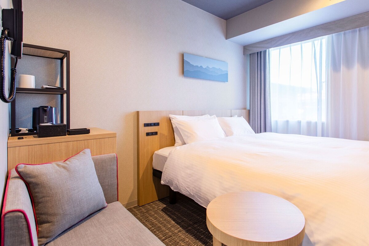 Ein Hotelzimmer mit einem Bett, einem Nachttisch mit Lampe, einem kleinen runden Tisch, einer Couch und einem Fenster mit Vorhängen.