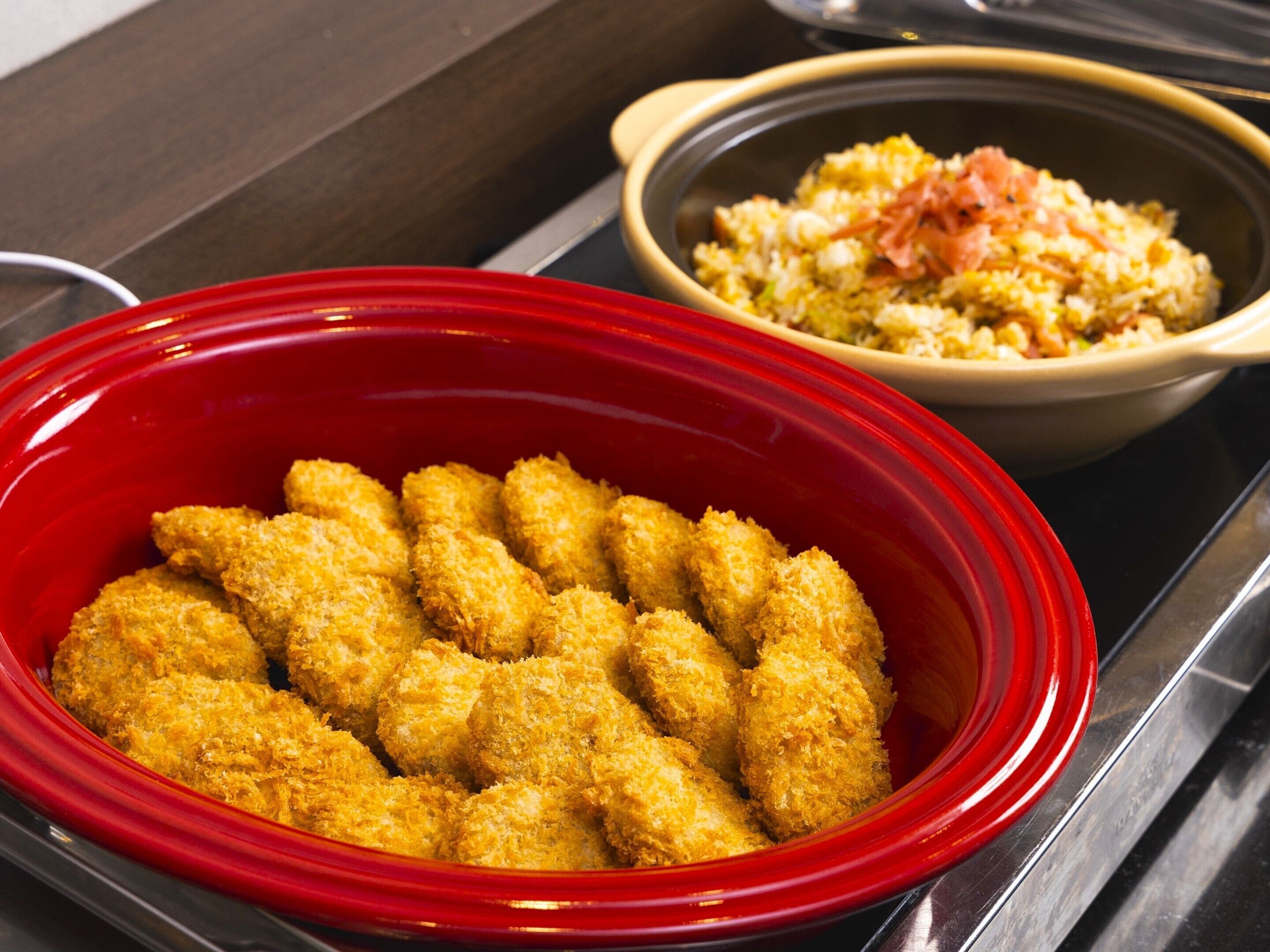 A red slow cooker filled with breaded chicken tenders and a bowl of rice with ham in the background.