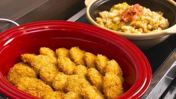 A red slow cooker filled with breaded chicken tenders and a bowl of rice with ham in the background.