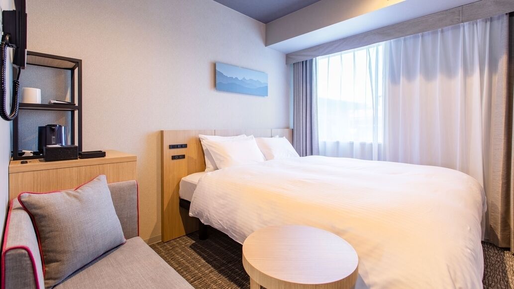 Ein Hotelzimmer mit Bett, Schreibtisch mit Telefon, Stuhl, kleinem runden Tisch und Fenster mit Vorhängen.