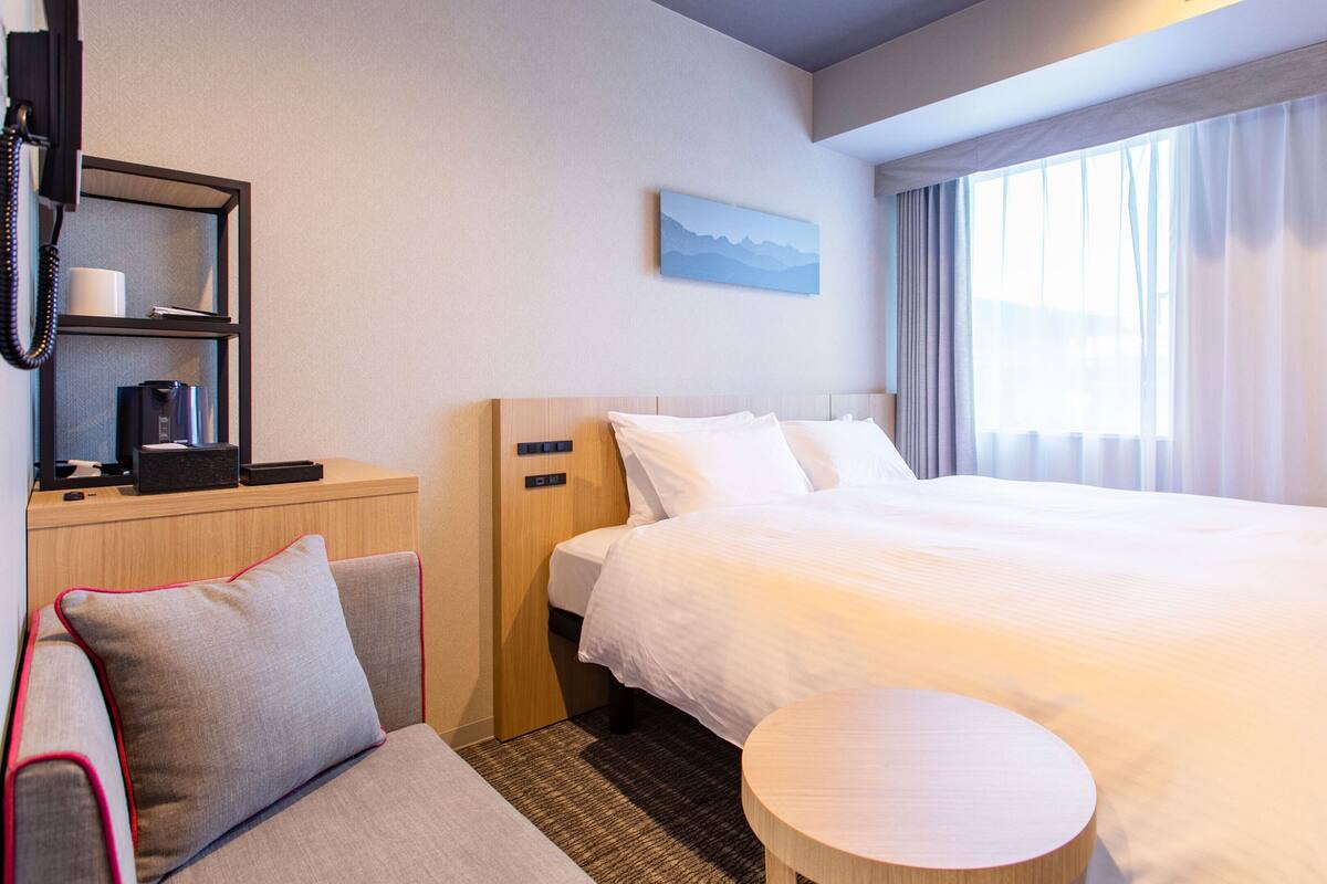 Een hotelkamer met een groot bed, een nachtkastje met een lamp, een klein rond tafeltje, een bank met kussens, een bureau met een computer en een koffiezetapparaat, en een schilderij aan de muur.