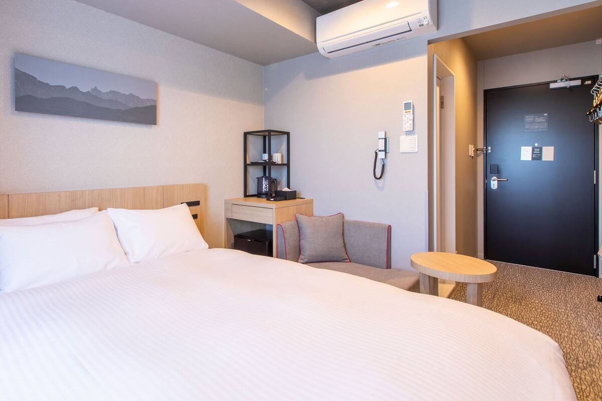 Een hotelkamer met een groot bed, een bureau met stoel, een kleine tafel, een airconditioningapparaat, een deur en een schilderij aan de muur.