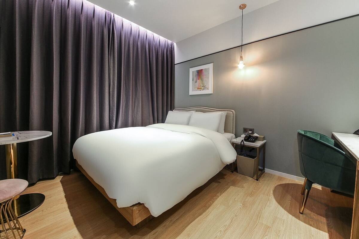 A modern hotel room with a large bed, bedside table, and a desk with a chair.