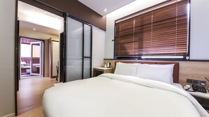 A modern hotel room with a large bed, a wooden headboard, a bedside table with a phone and charger, and a window with blinds. - Gijang No.25 Hotel (Busan)