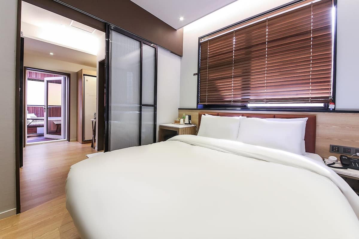 A modern hotel room with a large bed, a wooden headboard, a bedside table with a phone and charger, and a window with blinds.