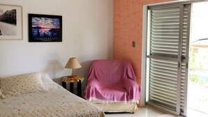 Un dormitorio con cama, ventilador de techo, una silla con manta rosa, mesita de noche con lámpara y ventana con persianas.