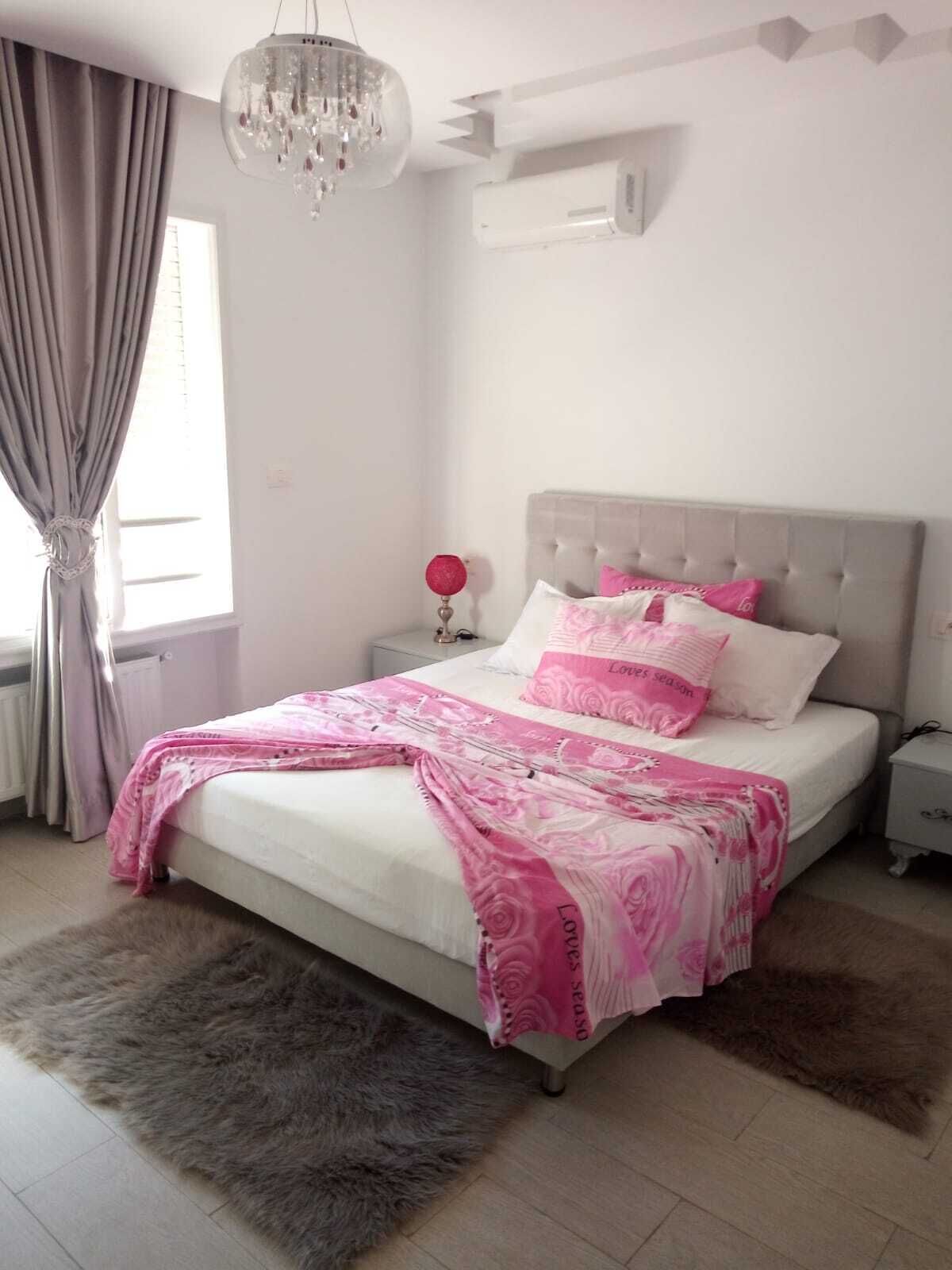 A bedroom with a bed, a nightstand, a chandelier, and a window with curtains.