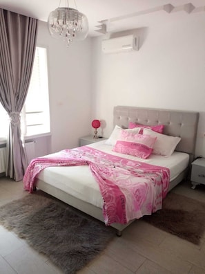 A bedroom with a bed, a nightstand, a chandelier, and a window with curtains.