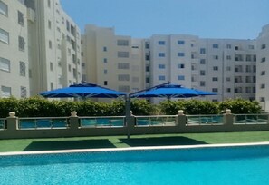 Una piscina con sombrillas azules, rodeada por setos bajos y una barandilla de metal.