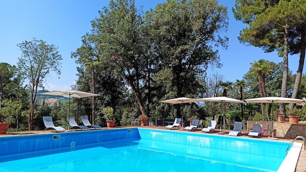 Ein Swimmingpool mit Liegestühlen und Sonnenschirmen, umgeben von Bäumen und blauem Himmel.