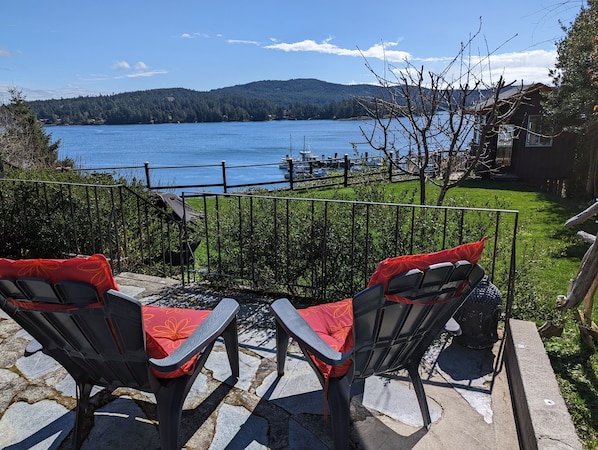 Un patio con dos sillas y una parrilla, con vista a un lago con barcos y una colina boscosa al fondo.