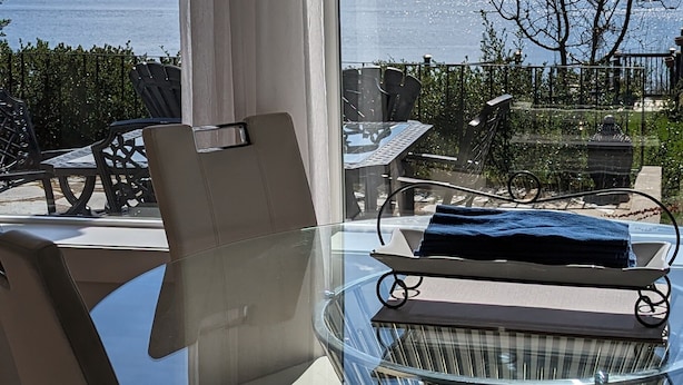 Une chambre moderne avec une table et des chaises en verre, une vue sur un lac et des montagnes, et un balcon aménagé avec du mobilier d’extérieur.