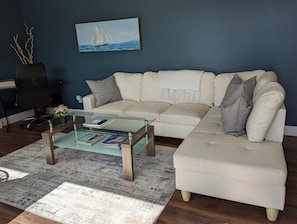Un salón con un sofá seccional color beige, una mesa de centro de vidrio, una pared azul oscuro y un cuadro de un velero.