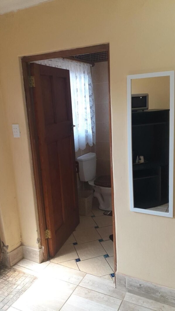 A room with a wooden door leading to a bathroom, a tiled floor, and a white wall with a small shelf. - Karl Heyne Guesthouse Double Room (Rustenburg)