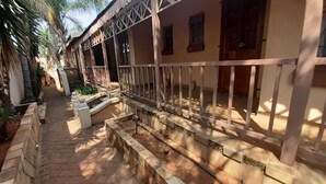 A covered outdoor area with a stone pathway leading to a building with a white railing and a wooden door. - Karl Heyne Guesthouse Double Room (Rustenburg)