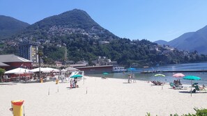 A beach with umbrellas, a lifeguard tower, and a mountain in the background. - Casa Hürlimann Erholung Im Malcantone (Alto Malcantone)