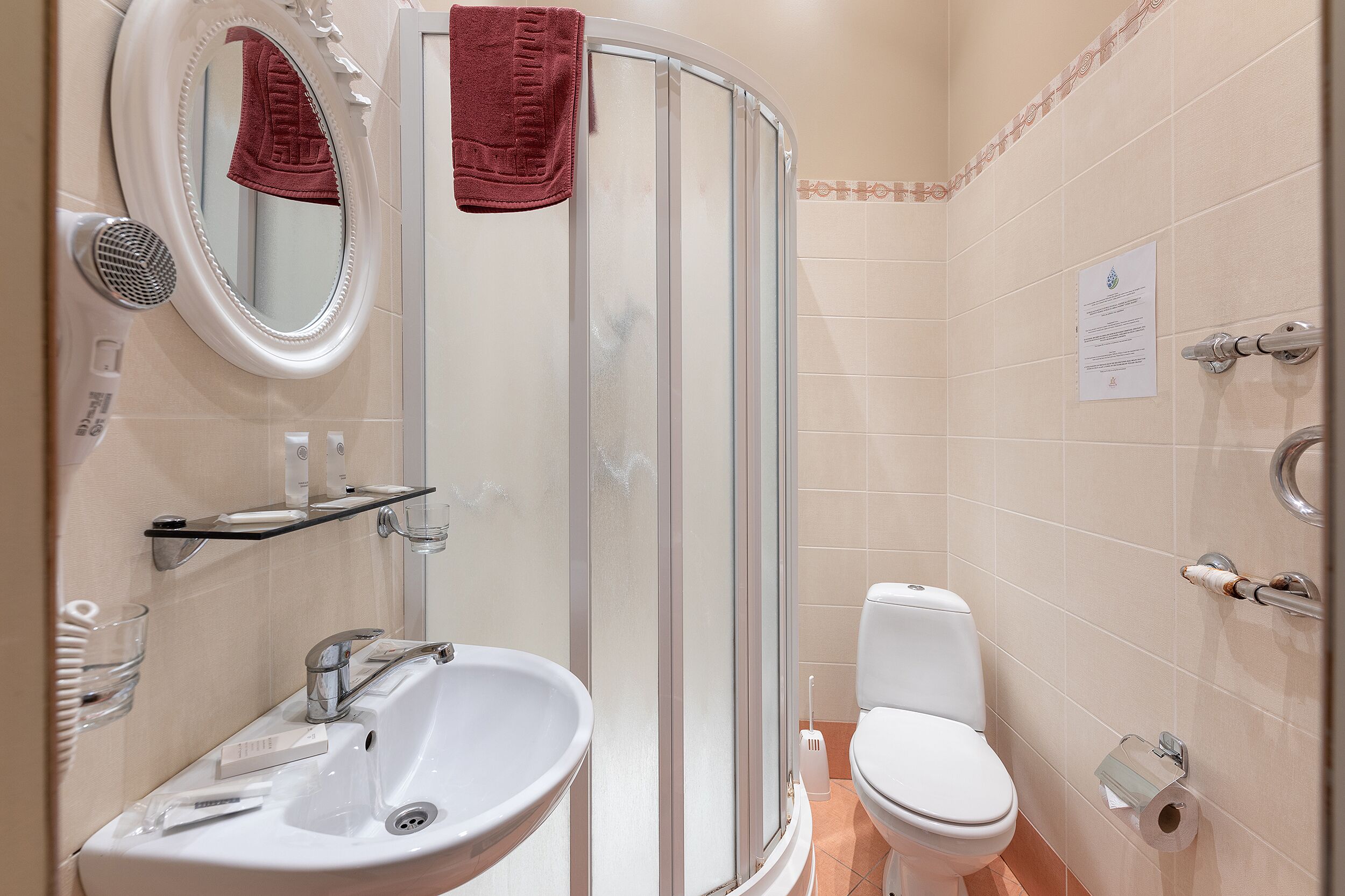 A bathroom with a sink, mirror, towel rack, shower, toilet, and a paper sign on the wall.